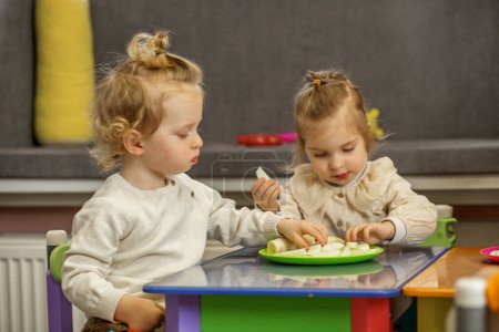 Zwei Kleinkinder teilen sich Snack auf buntem Spieltisch und unternehmen gemeinsam soziale und Lernaktivitäten.