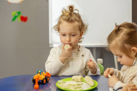 Mädchen isst während der Imbisszeit nachdenklich Banane am Spieltisch mit einem anderen Kleinkind in der Kita.