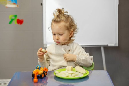 Preschool boy looks contemplatively at a piece of banana during snack break at colorful play table.