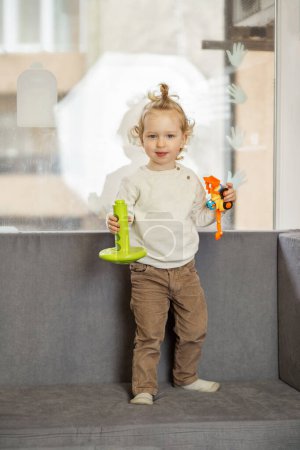 Little child stands on a couch holding a toy trumpet and figurine, looking playful and curious indoors.