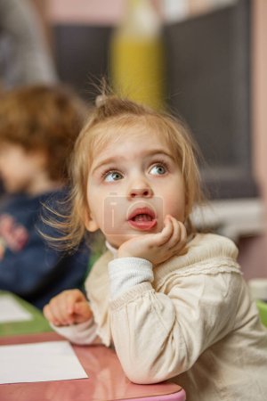 Little girl with big blue eyes looks thoughtful while participating in an activity in her preschool classroom.