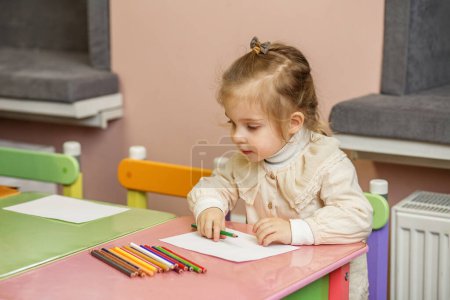 Kleines Mädchen mit Schleife im Haar konzentriert sich auf ihre Farbaktivitäten, sitzt am bunten Kindertisch.