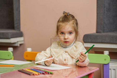Kleines Mädchen mit Schleife im Haar konzentriert sich auf ihre Farbaktivitäten, sitzt am bunten Kindertisch.