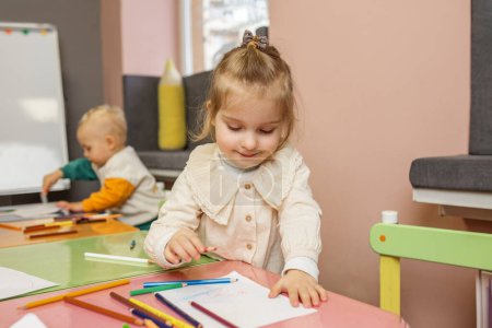Zufriedenes Kleinkind lächelt, als sie mit Buntstiften am rosafarbenen Tisch zeichnet, mit einem anderen Kind im Hintergrund.