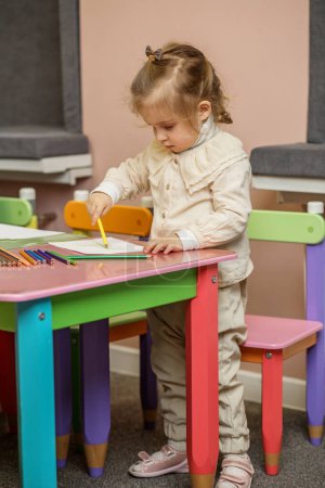 Focused toddler girl stands and draws intently at vibrant multi-colored children's table.