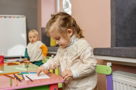 Contenu tout-petit fille sourit comme elle dessine avec des crayons de couleur à la table rose, avec un autre enfant en arrière-plan