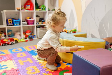 Toddler is sitting on colorful alphabet mat in playroom, focused on puzzle piece in thoughtful moment.