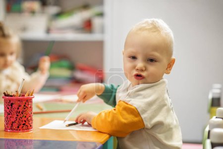 Little boy with focused expression draws at table in preschool classroom, with colorful pencils and another child nearby.