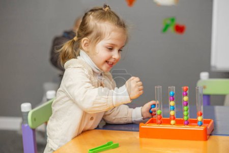 Smiling child girl with ponytail enjoys playful learning experience with colorful bead sorting toy in classroom.
