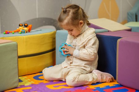 Little girl sits on colorful play mat, fully absorbed in solving handheld puzzle toy in preschool environment.