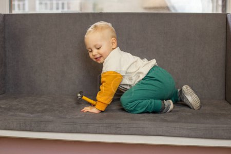 Alegre niño ríe mientras juega con el martillo de juguete en el sofá gris acogedor, que encarna la felicidad y la inocencia.