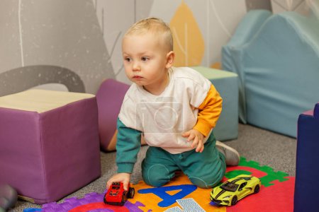Bébé concentré jouant avec des voitures de jouet rouges et jaunes sur un tapis de jeu multicolore, améliorant la coordination et les compétences de jeu.