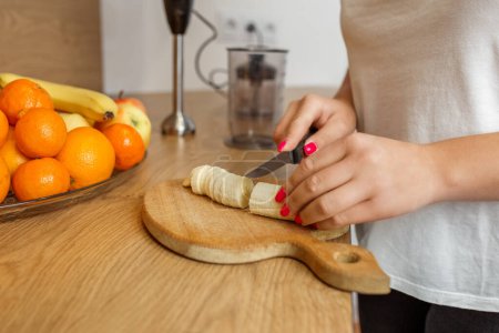 Vista de cerca de las manos de la mujer cortando plátano en una tabla de madera, con naranjas y licuadora en el fondo.