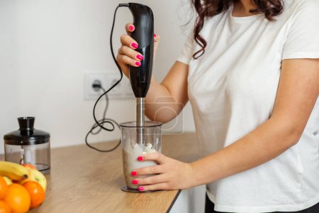 Woman in casual white shirt is using an immersion blender to mix banana and mascarpone smoothie.