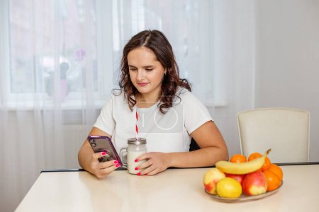Young girl is browsing her smartphone while enjoying a banana mascarpone smoothie at home with fresh fruits on table.