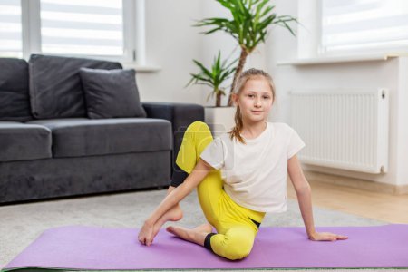 Téléchargez les photos : Jeune fille souriante dans une chemise blanche et leggings jaunes faire des exercices d'étirement sur un tapis de yoga violet à la maison. - en image libre de droit