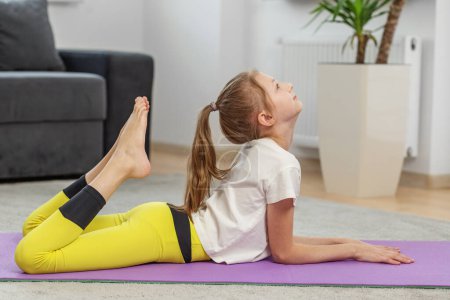 Téléchargez les photos : Jeune fille effectue un étirement de yoga complexe, levant le pied vers sa tête sur le tapis violet, montrant flexibilité et compétence. - en image libre de droit