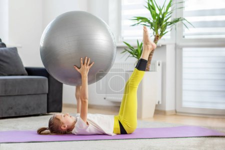 Kindermädchen liegt zu Hause auf einer Yogamatte, hebt mit Beinen und Armen einen Fitnessball und macht eine Kernkraftübung.