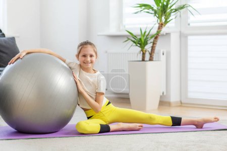 Lächelndes Teenie-Mädchen mit silbernem Fitnessball sitzt auf einer lila Yogamatte und genießt ihr Workout im sonnigen Raum.