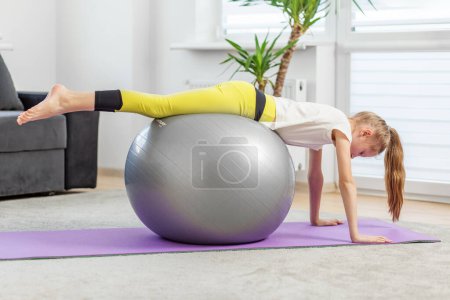 Konzentriertes Mädchen behält Plankenstellung auf Fitnessball bei, arbeitet an ihrem Kern und balanciert zu Hause auf lila Yogamatte.