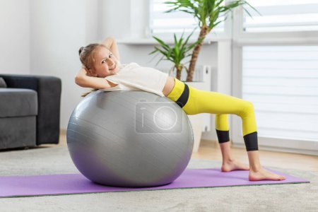 Junges Mädchen macht Backbend auf Fitnessball und entspannt, während sie zu Hause ihre Rückenmuskeln auf einer lila Yogamatte dehnt.