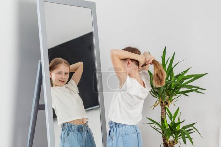 Child ties her hair into ponytail while observing herself in mirror in well-lit room with green plant.