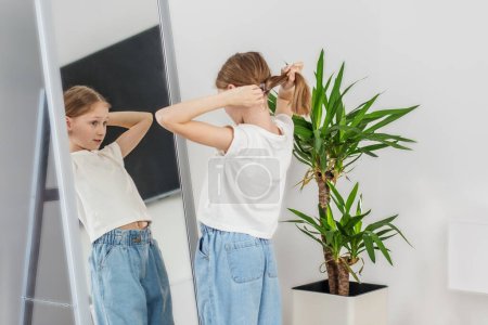 Preteen child ties her hair into ponytail while observing herself in mirror in well-lit room with green plant.