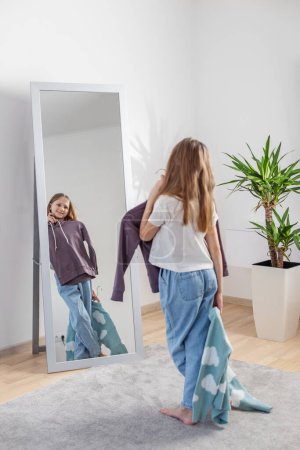Mädchen untersucht ihre Outfit-Optionen, die sich in einem Ganzkörperspiegel widerspiegeln, umgeben von einem gemütlichen Ambiente mit Zimmerpflanze.