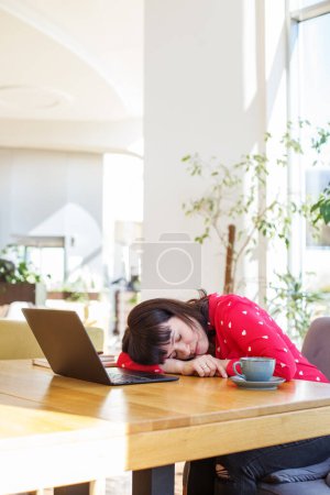Erschöpfte junge Geschäftsfrau schläft am Schreibtisch mit Laptop und Tasse Kaffee im sonnenbeschienenen, pflanzengeschmückten Büro.