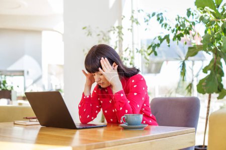 Worried professional woman feeling stressed while working on her laptop at bright office desk with coffee cup.