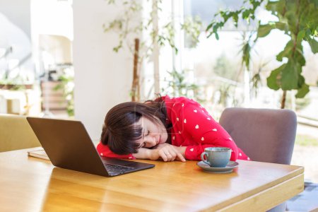 Erschöpfte Millennial-Geschäftsfrau schläft mit Laptop und Tasse Kaffee am Schreibtisch im sonnenbeschienenen, pflanzengeschmückten Büro.