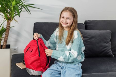 Cheerful preteen child smiling while packing red backpack, sitting comfortably on dark sofa at home.