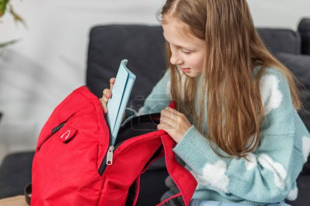Photo for Focused preteen girl sitting on couch, looking inside her open red backpack, with book beside her. - Royalty Free Image