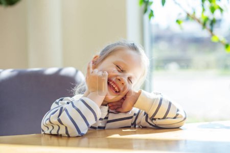 Cheerful young girl laughing heartily while sitting at a wooden table bathed in sunlight.
