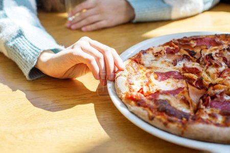 Person's hand reaching to grab slice of meat lover's pizza with crispy bacon and melted cheese on sunny table.