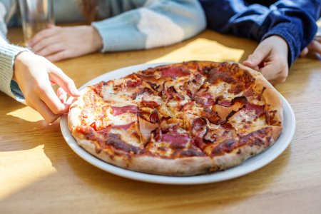 Family hands reaching to take slices of delicious meat lover's pizza on sunny wooden table, sharing meal together.