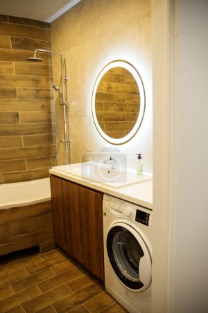 Stilvolles Badezimmer mit hölzerner Akzentwand, rund beleuchtetem Spiegel und integrierter Waschmaschine unter eleganter Arbeitsplatte.