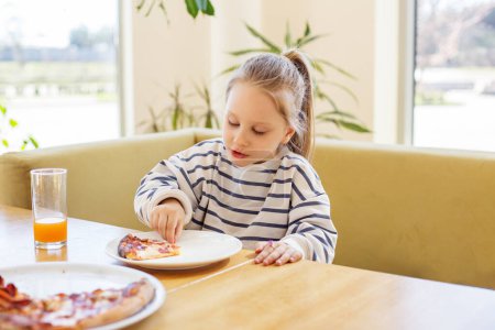 Petit enfant manger intensément tranche de pizza tout en étant assis à la table intérieure ensoleillée avec un verre de jus d'orange.