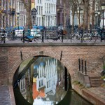 Bare trees without leaves in winter and historic facades and Dutch bikes on the bridge with classic lampposts, above the yards of a canal with brick walls and stairs and doors, reflecting in the water