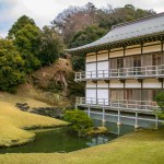A traditional wooden structure with a white facade and tiled roof stands at Kenchoji temple in Kamakura, Japan. A bridge connects the building to a landscaped bonsai garden beside a tranquil pond.