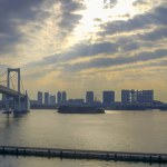 View capturing the Rainbow Bridge arching over Tokyo Bay toward Odaiba, the man-made island, against a golden sky at sunrise. The silhouette of Tokyo cityscape lines the horizon, while boats navigate