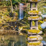 A serene scene from a Japanese garden. A stone pagoda, partially covered in snow and moss, stands by a tranquil pond. In the backdrop, a waterfall gently cascades amidst lush green foliage. The