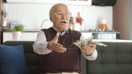 Téléchargez les photos : Heureux vieil homme excité par ses dollars. Le vieil homme est assis sur le canapé et compte les billets. Portrait d'une personne heureuse parce qu'il gagne beaucoup d'argent en pariant. - en image libre de droit
