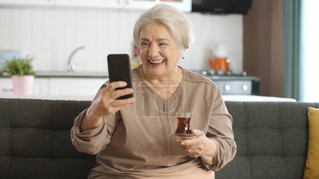 Téléchargez les photos : Bonne vieille grand-mère tenant téléphone portable, appel vidéo avec les proches tout en buvant du thé. Une femme âgée apprenant à utiliser un appareil technologique moderne. Utilisation de la technologie chez les personnes âgées. - en image libre de droit
