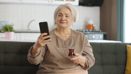 Téléchargez les photos : Heureuse vieille grand-mère tenant téléphone portable est très surprise d'entendre qu'elle fait appel vidéo avec ses proches tout en buvant du thé. Une femme âgée apprenant à utiliser un dispositif technologique moderne. - en image libre de droit