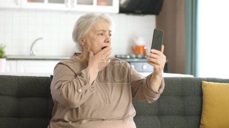 Téléchargez les photos : Femme âgée faisant un appel vidéo avec des parents, regardant la caméra et saluant, assis sur un canapé confortable. Concept d'utilisation de la technologie avec les aînés. - en image libre de droit