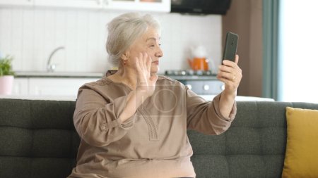 Téléchargez les photos : Femme âgée faisant un appel vidéo avec des parents, regardant la caméra et saluant, assis sur un canapé confortable. Concept d'utilisation de la technologie avec les aînés. - en image libre de droit