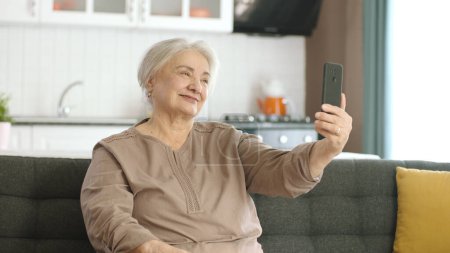 Seniorin lächelt, telefoniert per Video mit Verwandten, schaut in die Kamera und winkt, sitzt auf bequemem Sofa. Technologienutzungskonzept mit Senioren.