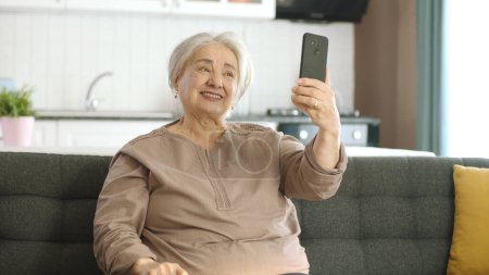 Téléchargez les photos : Femme âgée souriant, faire un appel vidéo avec la famille, regardant la caméra et agitant, assis sur un canapé confortable. Concept d'utilisation de la technologie avec les personnes âgées. - en image libre de droit