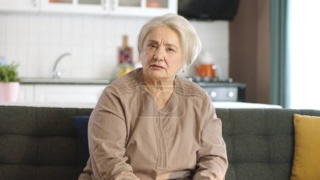 Una mujer mayor con el pelo blanco está discutiendo con la otra persona en una videollamada en línea. Mujer mayor enojada con problemas haciendo videollamada en el portátil en el sofá en la sala de estar en casa. Vista de cámara frontal.